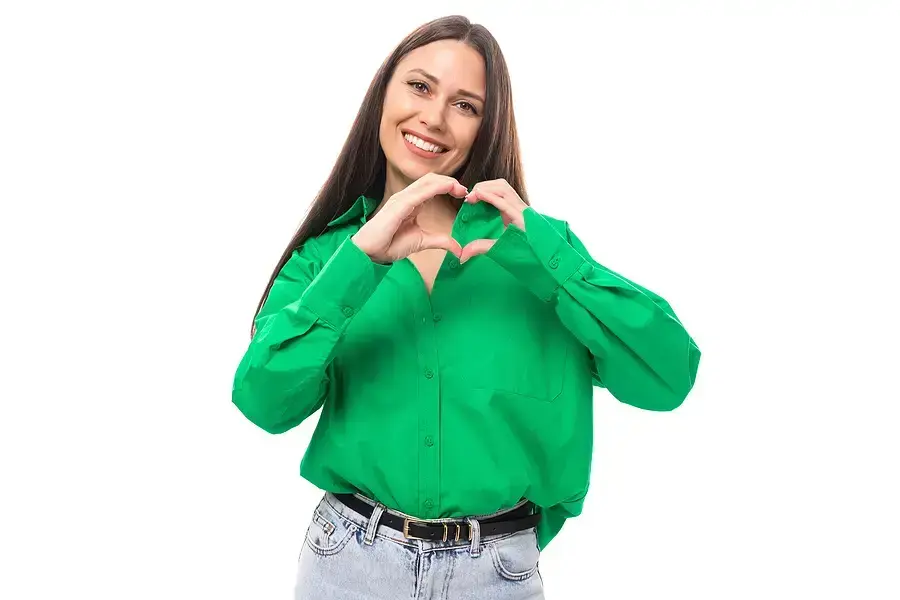 Happy woman in green shirt making heart hand gesture promoting dental health and wellness.