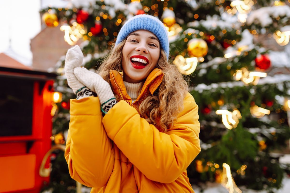 Happy woman in yellow jacket smiling at Christmas market with decorated tree backdrop.